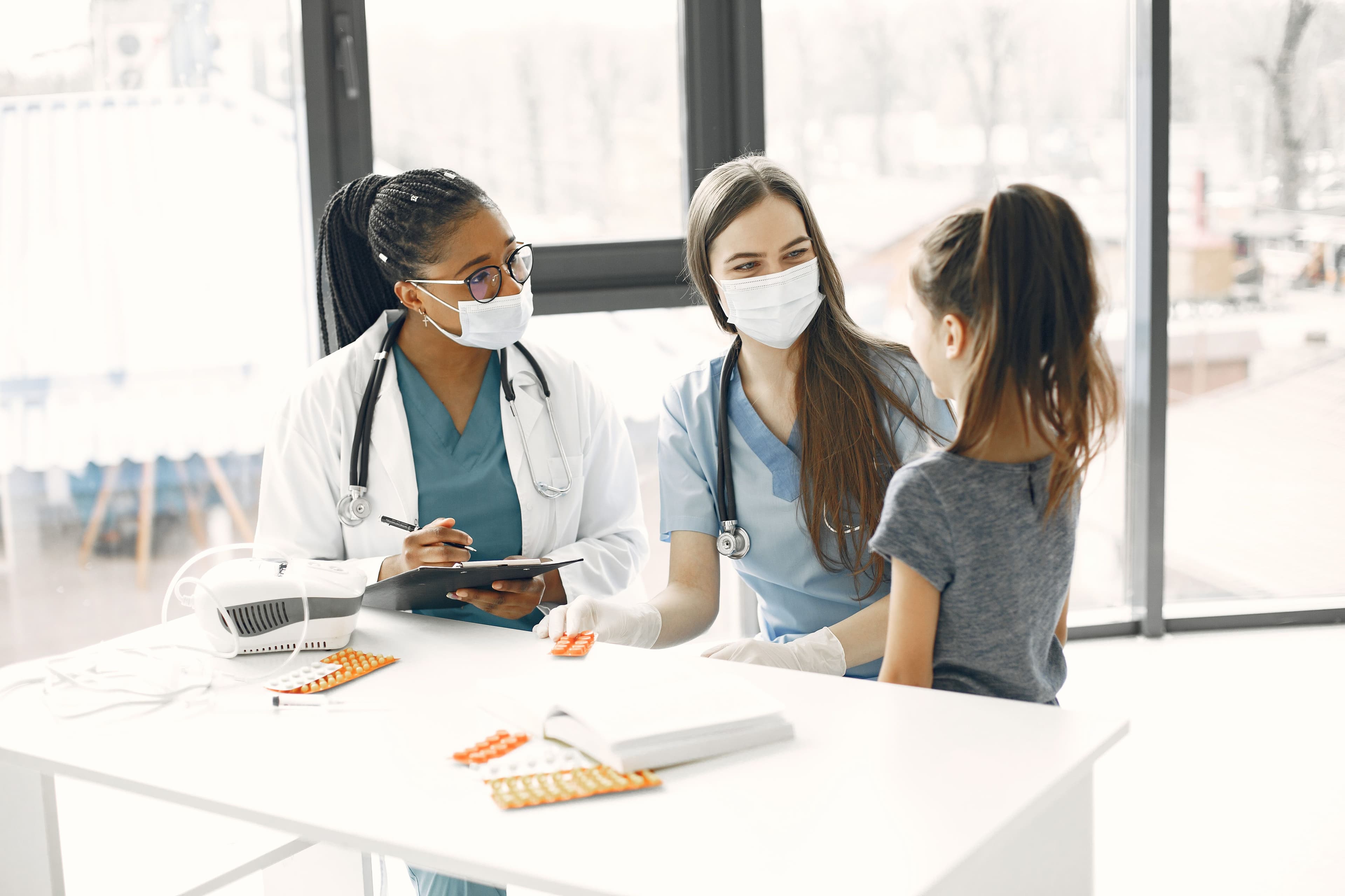 Una doctora y una enfermera con mascarillas atienden a una niña en un consultorio médico.